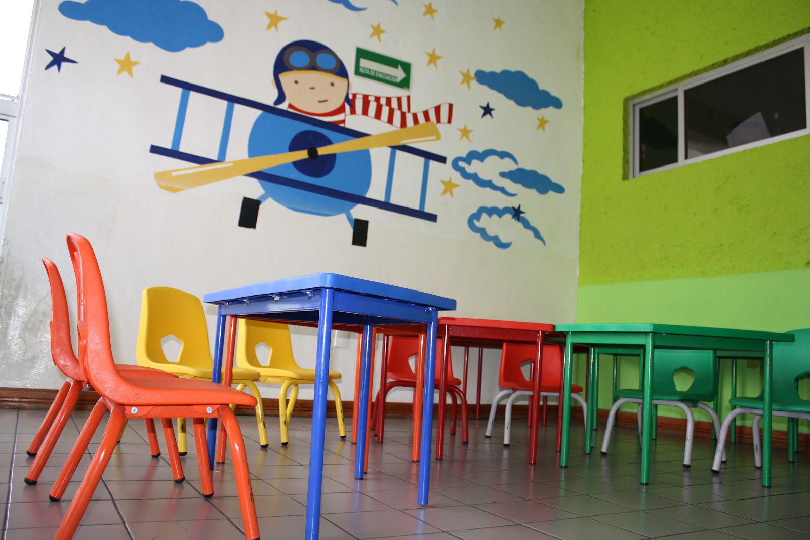 Salón de Bügu Guardería con mesitas y sillas de colores y mural de avión